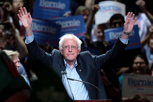 Bernie Sanders raising his arms above his head to salute the crowd at a political rally.